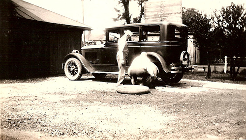 A black and white photo of a horse and a car with a flat tire.