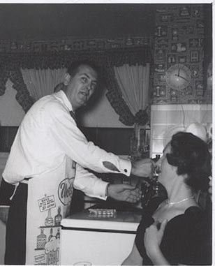 A man learning to cook, wearing an apron as he feeds a woman in a kitchen.