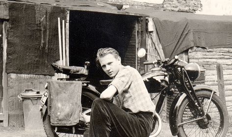 A man sitting on a motorcycle, his first motorcycle, in front of a shack.