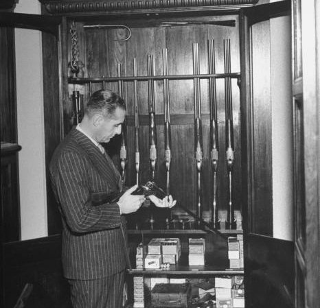 A man in a suit standing in front of a gun cabinet, showcasing his home defense arsenal.