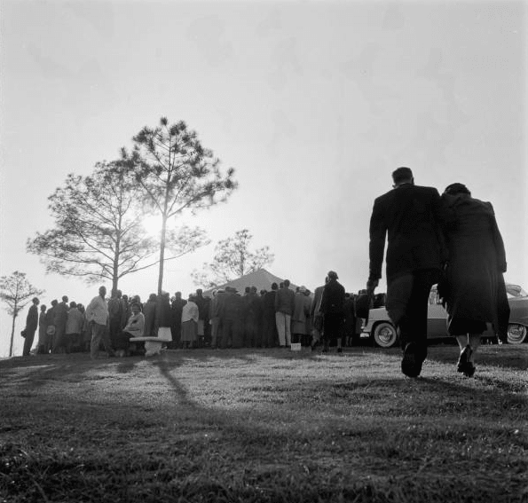 A black and white photograph of a crowd of people walking in the grass, with a man following funeral etiquette.