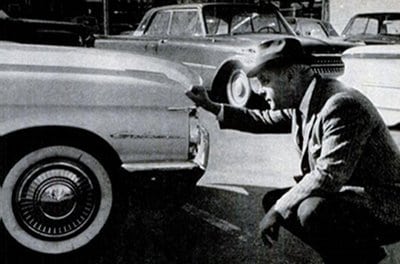 A man kneeling down next to a parked car, contemplating whether to buy the used car.