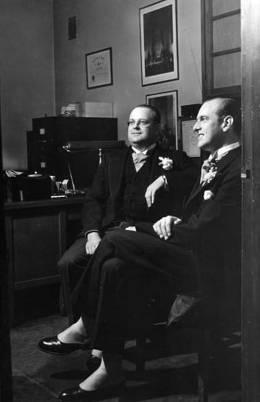 Two best men in suits discussing their duties in a room.