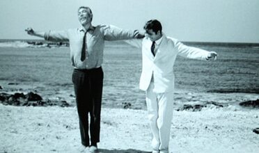 Two men performing a wild dance on a beach with their arms outstretched, reminiscent of Zorba the Greek.