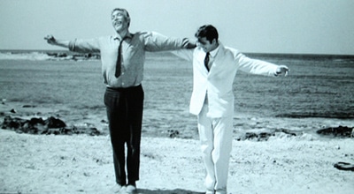 Two men performing a wild dance on a beach with their arms outstretched, reminiscent of Zorba the Greek.