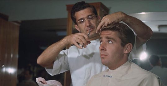 The man is getting a haircut while talking to the barber in a barbershop.