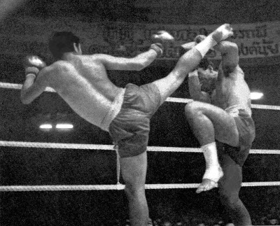 Two men engaging in Muay Thai, Thailand's National Sport, in a boxing ring and kicking each other.