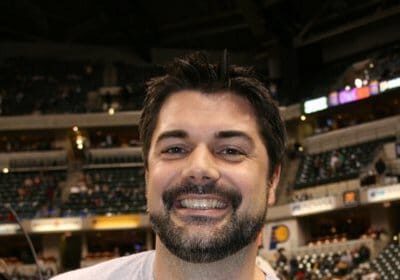 A man smiling in front of a basketball court, likely an NBA Strength and Conditioning Coach.