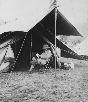 A manly man sitting in front of a tent reading a book.