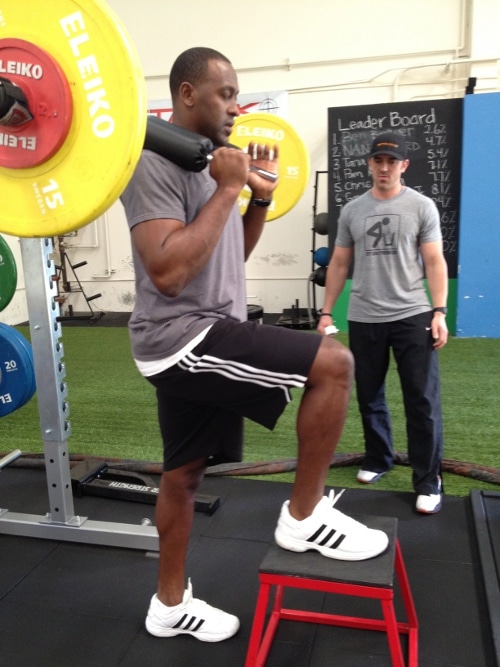 Two men standing in a gym, demonstrating squats and barbells. The gym owner provides insight into their job responsibilities and experiences.