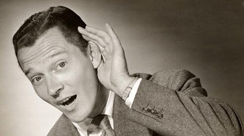 A black and white photo of a man paying attention, with his hand on his ear, actively listening and learning.