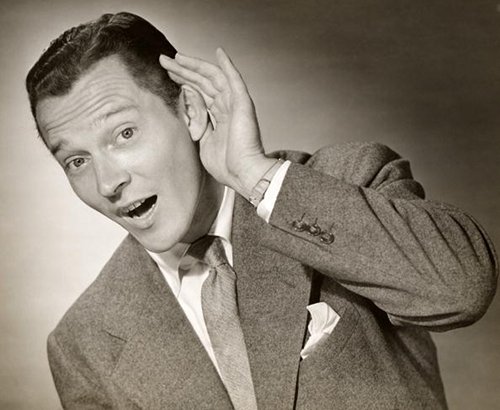 A black and white photo of a man paying attention, with his hand on his ear, actively listening and learning.