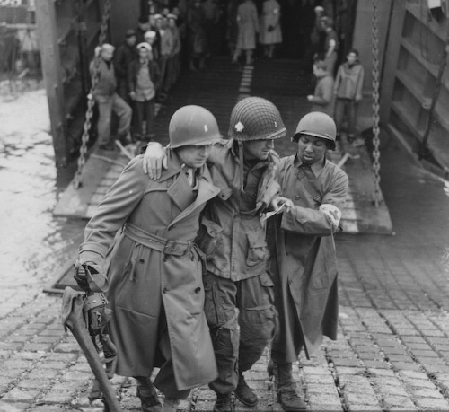A group of soldiers, led by a combat medic, are bravely walking on a bridge amidst the chaos of battle. Equipped to save lives, the medic remains vigilant for any signs of