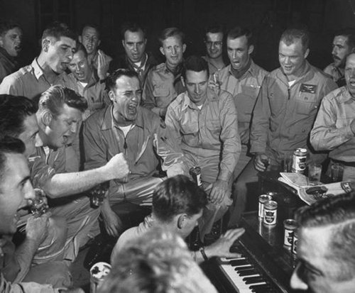 A round of men sitting around a piano, enjoying drinks.