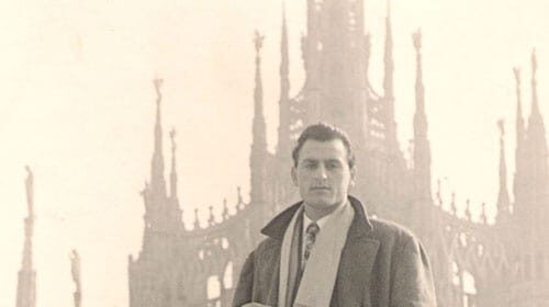 A man's guide, dressed in an overcoat, standing in front of a building.