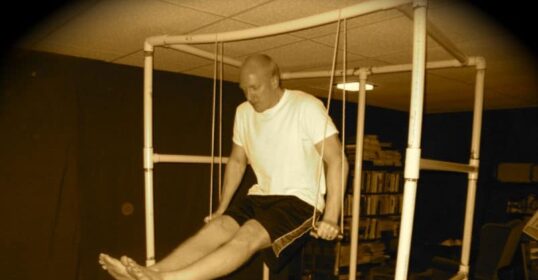 A man is using a PVC Pipe as part of his leg stretch routine in a room at his Ultimate Bodyweight Gym.