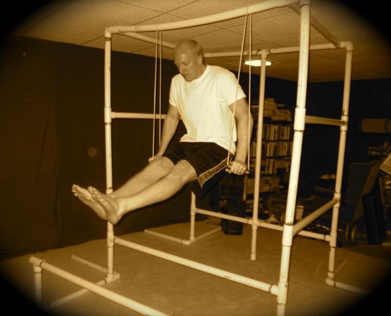 A man is using a PVC Pipe as part of his leg stretch routine in a room at his Ultimate Bodyweight Gym.