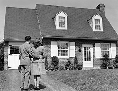 A black and white photo of a couple standing in front of their first home.