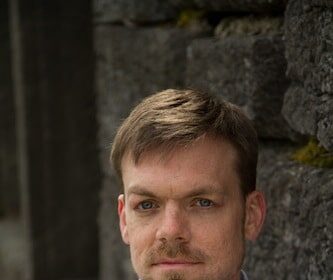 A So You Want My Job novelist in a blue shirt leaning against a stone wall.