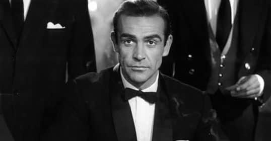 A man in a tuxedo with a bow tie sitting at a black-tie event, surrounded by others in formal attire.
