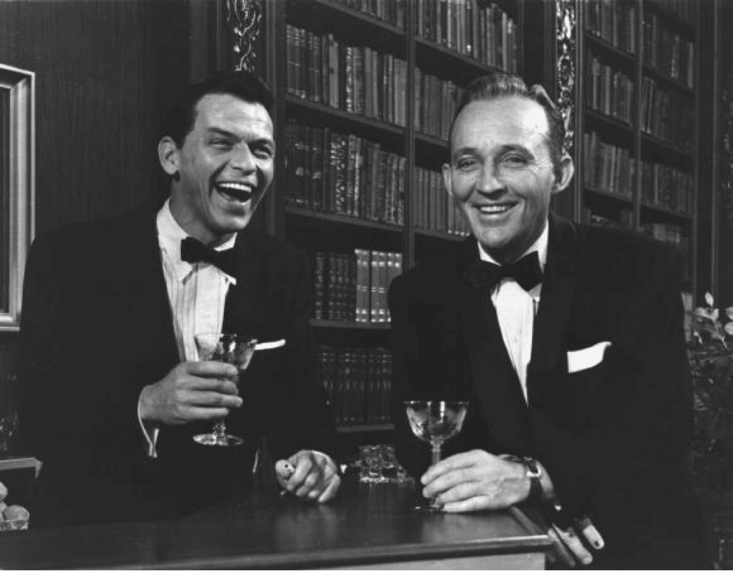 Two men in tuxedos smiling at each other, enjoying gin cocktails.