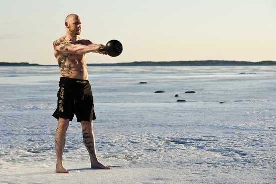 A man with tattoos showcasing his power swings a kettlebell in the snow.