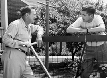 Two men standing next to a fence talking to each other.