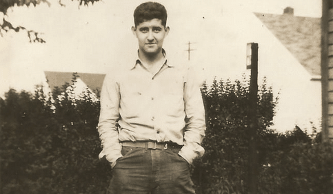 A young man with his hands in his pockets stands in front of a house in this old photo.