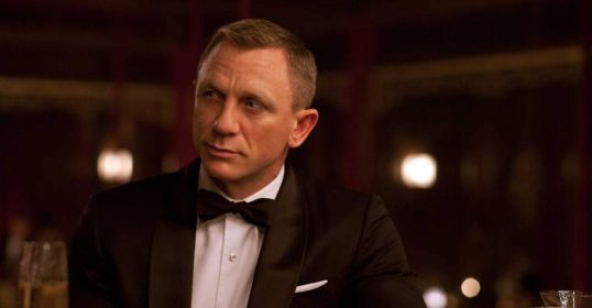 James Bond, with his suave savoir-faire, sits at a table in his iconic tuxedo.