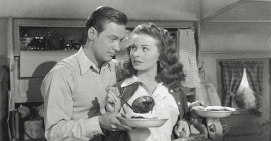 A man and woman, married, standing in a kitchen with a plate of food.