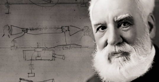 A man with a white beard standing in front of a drawing.