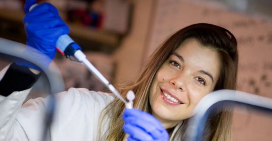 A woman in a lab coat holding a pipette while researching Genetics.