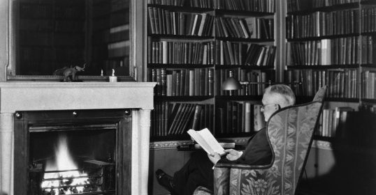 A man reading a book in front of a fireplace.