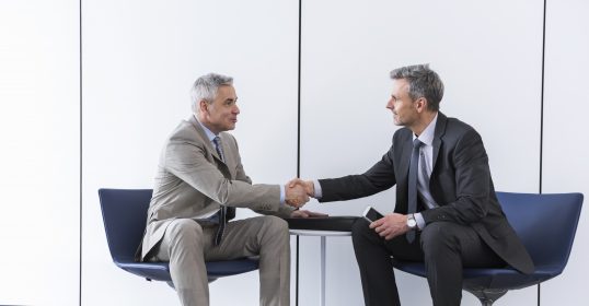 Two businessmen in formal dress shaking hands during negotiations in an office.