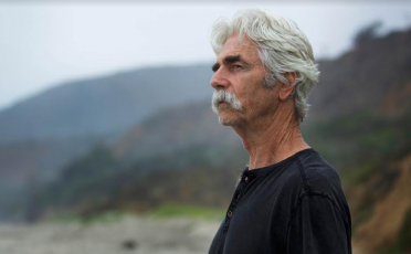 A man with a mustache, going gray, stands on a beach.
