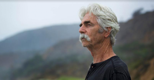 A man with a mustache, going gray, stands on a beach.