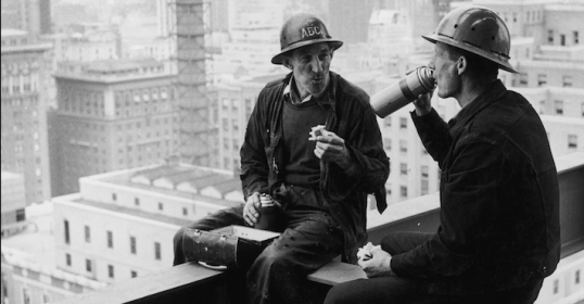 Two construction workers conversing on a ledge overlooking the city.