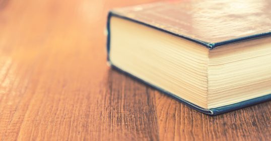 A long hardcover book lies closed on a wooden surface, its pages and spine hinting at the depth within.