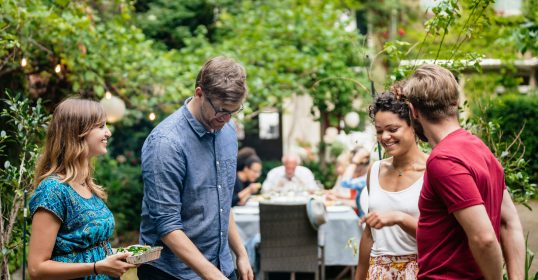 A group of people standing around a BBQ grill in a garden, having conversations.