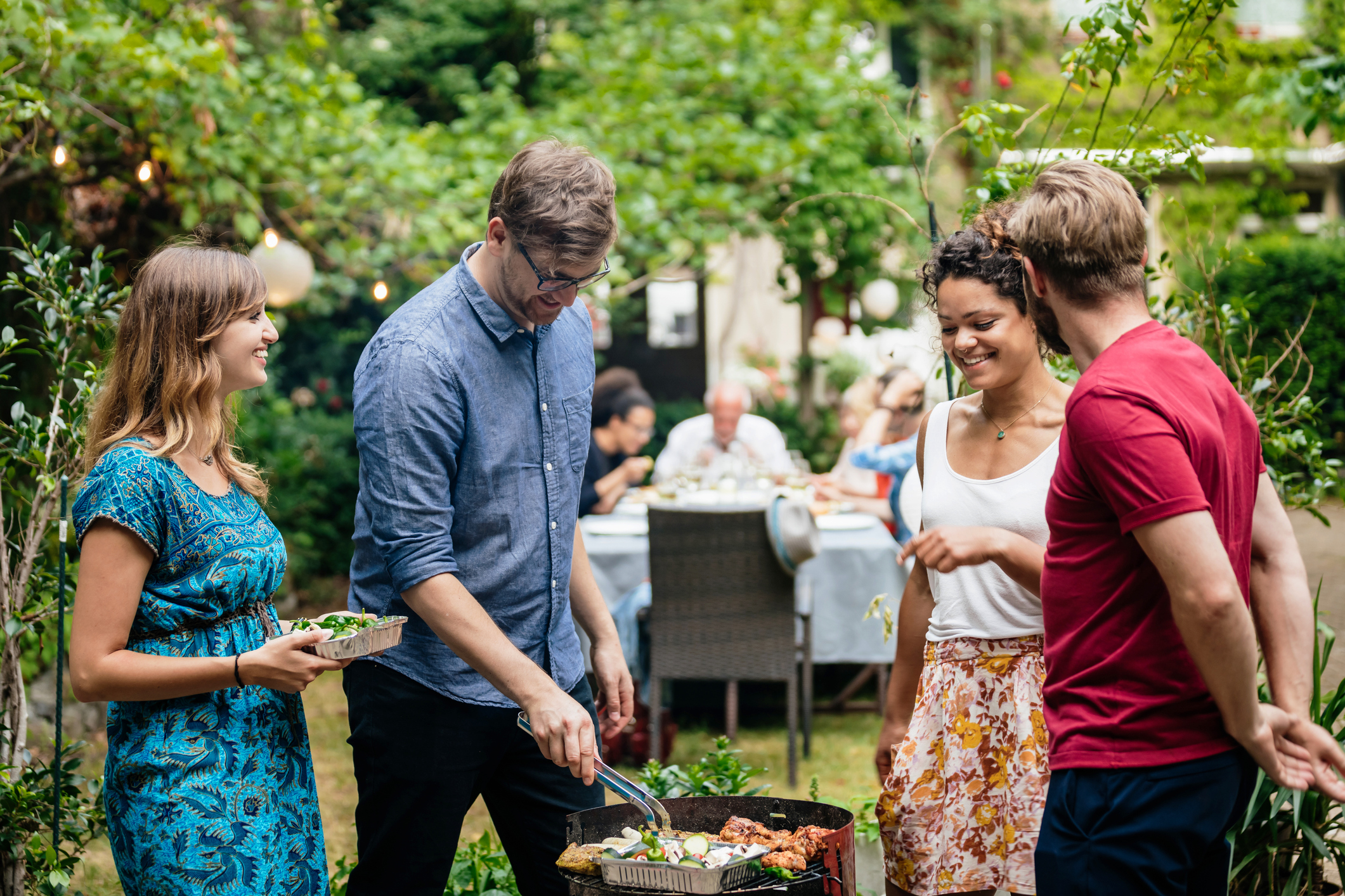 A group of people standing around a BBQ grill in a garden, having conversations.