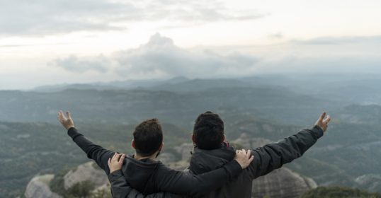 Two friends standing on top of a mountain, arms outstretched.