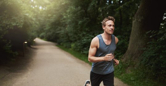 A man is jogging on a path in the woods, keeping his body parts active and relaxed.