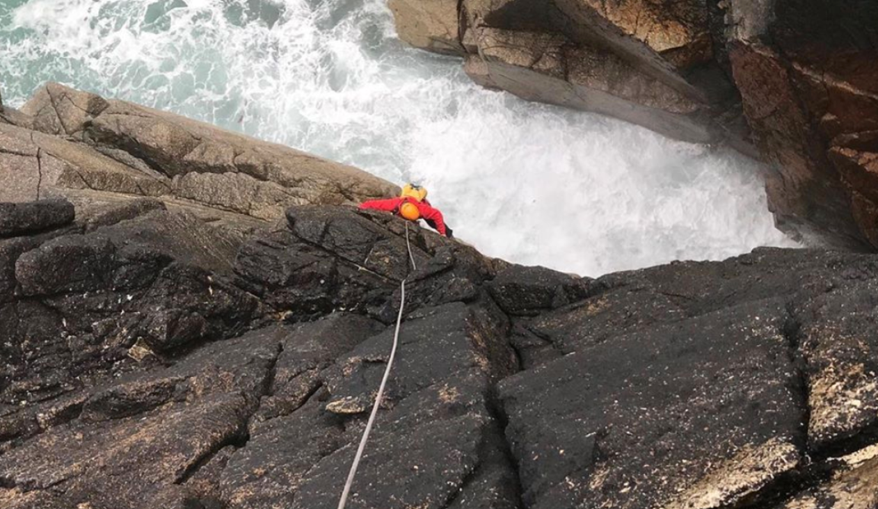 A man is climbing up a rocky cliff near the ocean, feeling uncomfortable yet determined.
