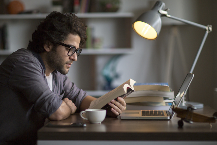 A man is immersed in a book at a desk with his laptop.