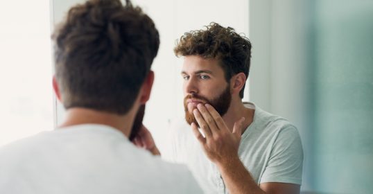 A man is brushing his beard in front of the mirror while pressing his beard oil.