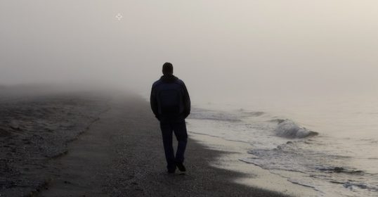 A lonely man stands on a foggy beach.