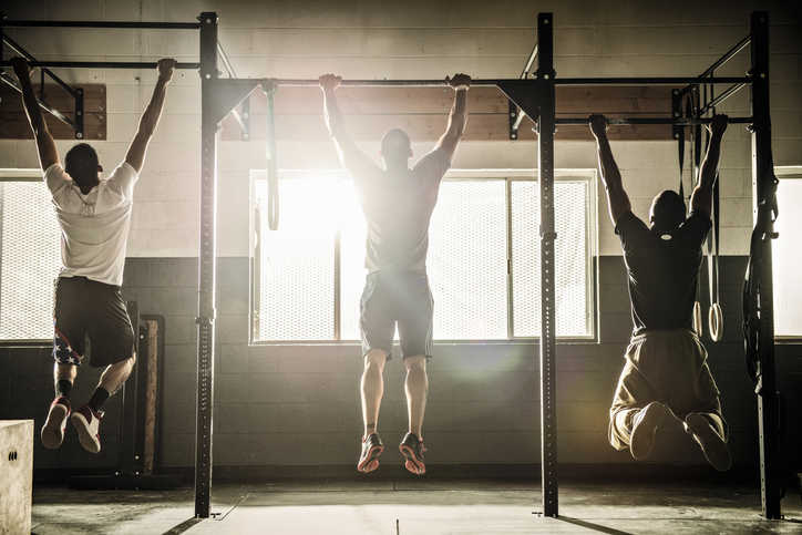 A group of men doing pull ups in a gym to reach their physical benchmarks.