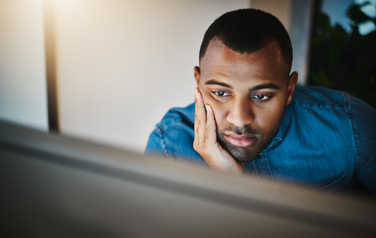 A man, bored, staring at a computer screen with his hand on his face.