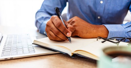 A man's hand writing in a notebook next to a laptop.