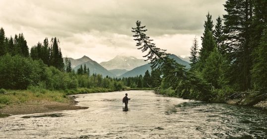 A man is fly fishing in a river with mountains in the background, reflecting on life lessons.
